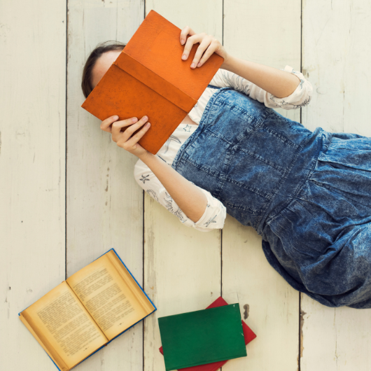 Femme allongée lisant un livre, entourée d'autres ouvrages pour enrichir son carnet de lecture.