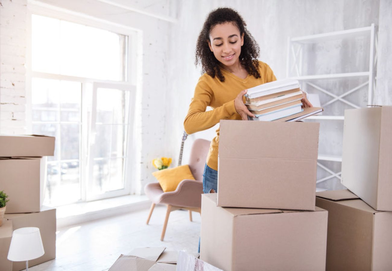 Favorite books. Pleasant curly-haired girl packing a pile of books into a big box before moving out of the room in the dormitory